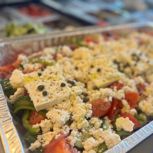A Greek salad with tomatoes, green peppers, feta cheese, capers, and olive oil in a foil tray.