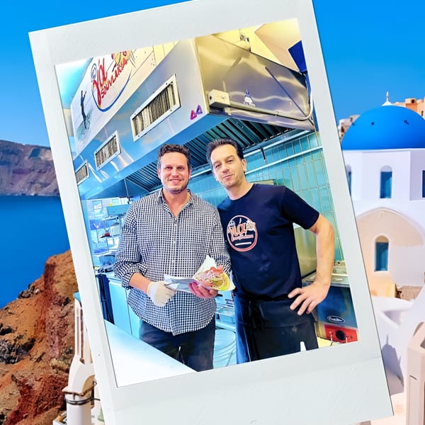 Two men in a kitchen, one holding a souvlaki, superimposed on a Greek island scene.