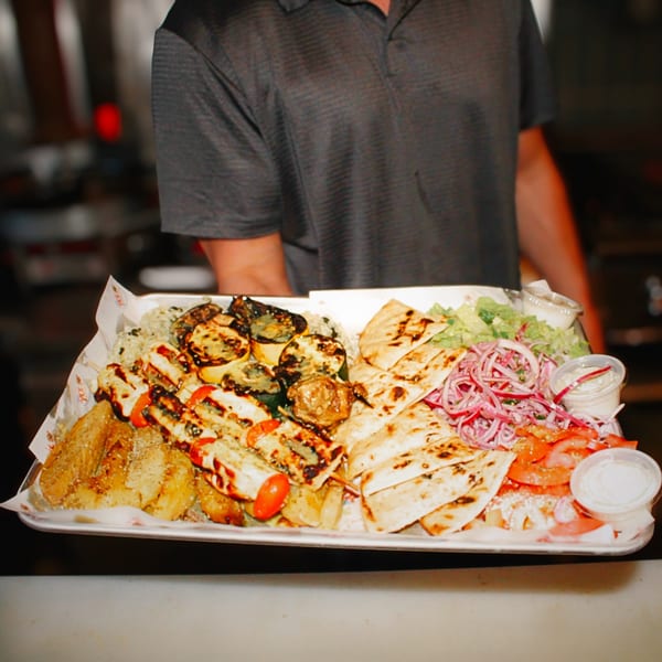A server holds a large tray of Greek food, including grilled meats, pita, potatoes, and salad.