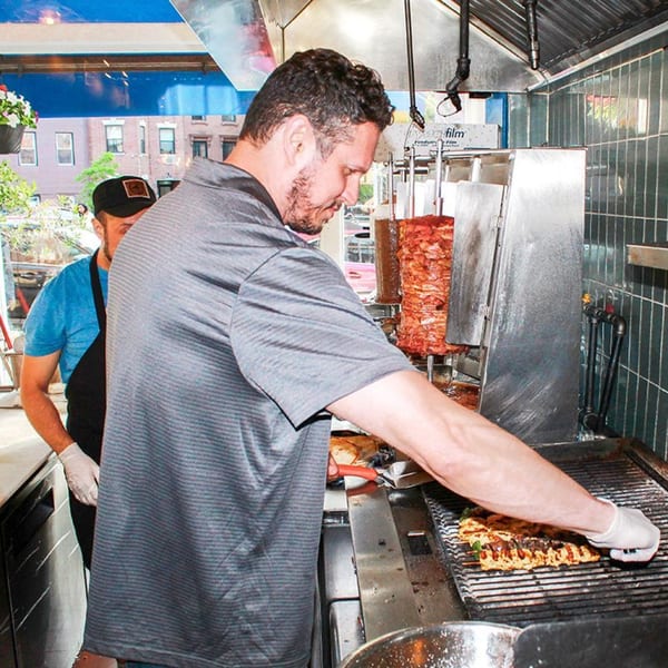 Two chefs grilling souvlaki skewers and carving meat from a vertical rotisserie at Let's Go Ya Souvlakia.
