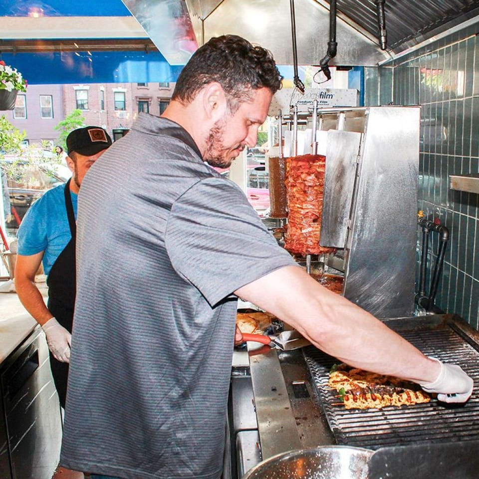 Two chefs grilling souvlaki skewers and carving meat from a vertical rotisserie at Let's Go Ya Souvlakia.