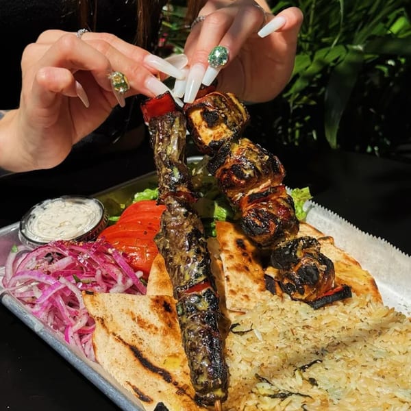 Hands with long, jeweled nails holding a Greek souvlaki skewer over a platter of food.