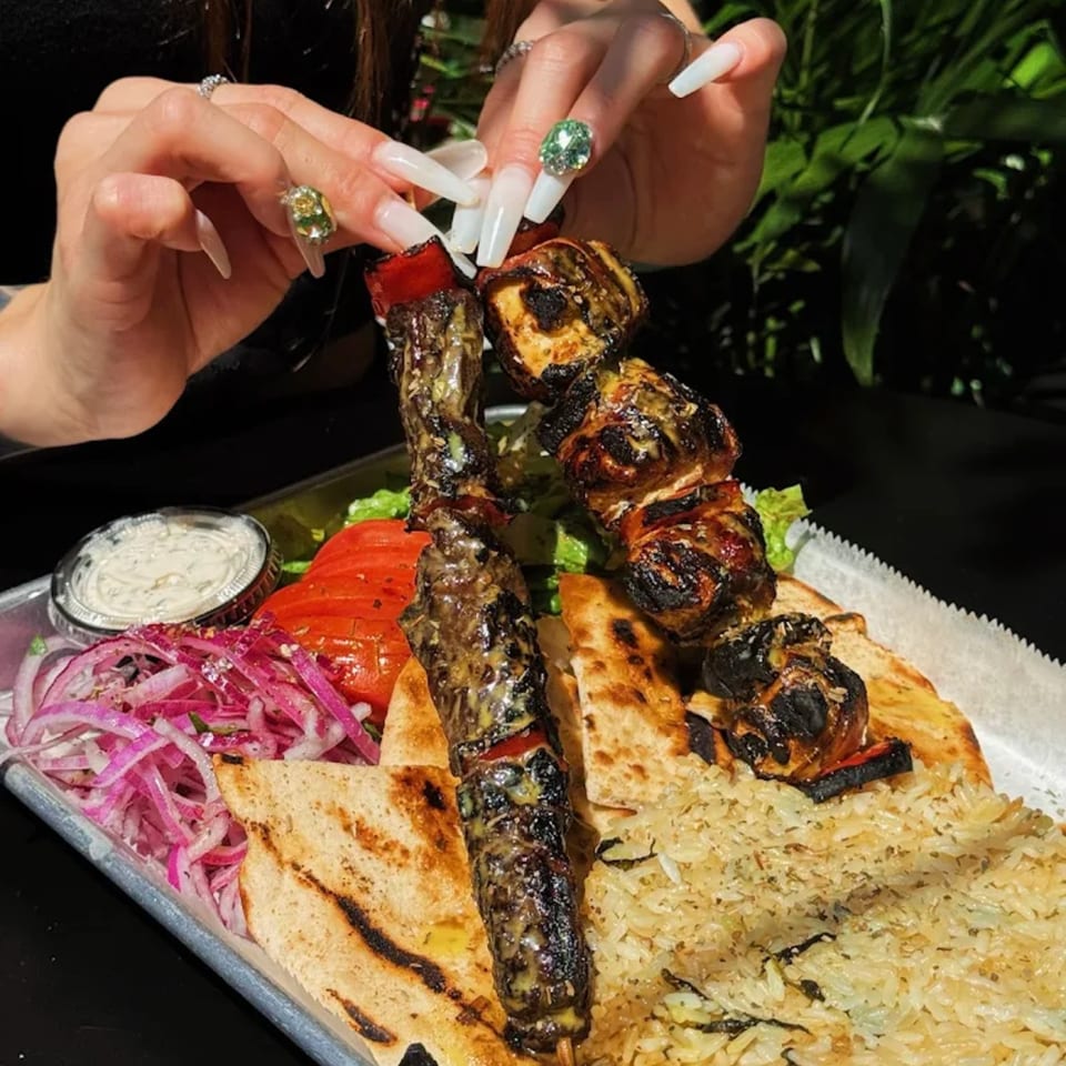 Hands with long, jeweled nails holding a Greek souvlaki skewer over a platter of food.