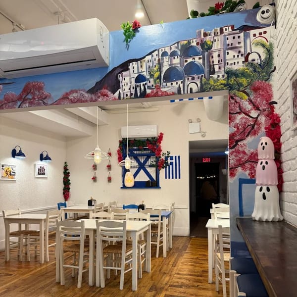 The interior of Let's Go Ya Souvlakia, featuring white tables and chairs, and a Greek-themed mural.