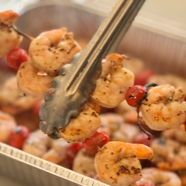 Tongs lifting a shrimp and cherry tomato skewer from a foil tray of Greek souvlaki.
