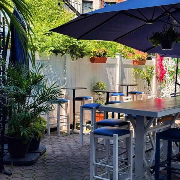 Outdoor patio of Let's Go Ya Souvlakia with blue umbrellas, high-top tables, and stools.